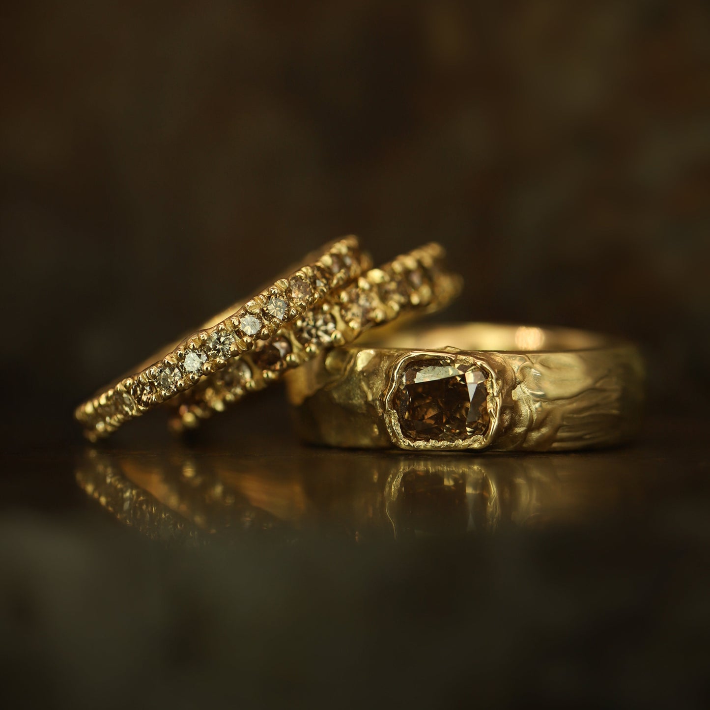Two gold rings displayed on a dark reflective surface. One ring features a large square brown gemstone, while the other has a row of small clear stones along its band.