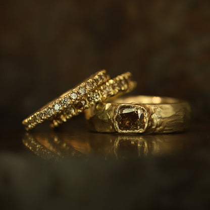 Two gold rings displayed on a dark reflective surface. One ring features a large square brown gemstone, while the other has a row of small clear stones along its band.