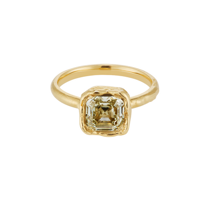 A gold ring featuring a square-cut, emerald-shaped clear gemstone in a textured bezel setting, shown on a plain white background.
