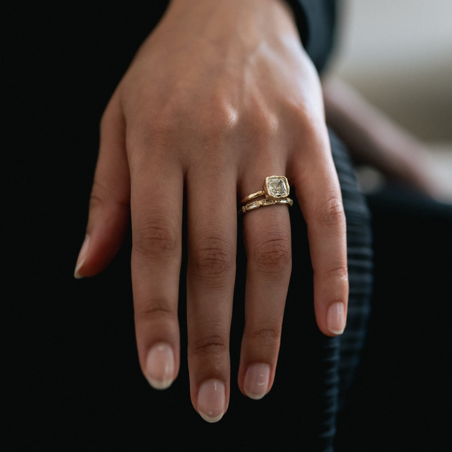 A close-up of a person's hand wearing a gold ring with a square-cut clear gemstone, set against a dark background.