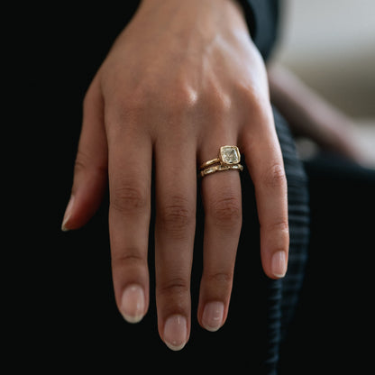 A close-up of a person's hand wearing a gold ring with a square-cut clear gemstone, set against a dark background.