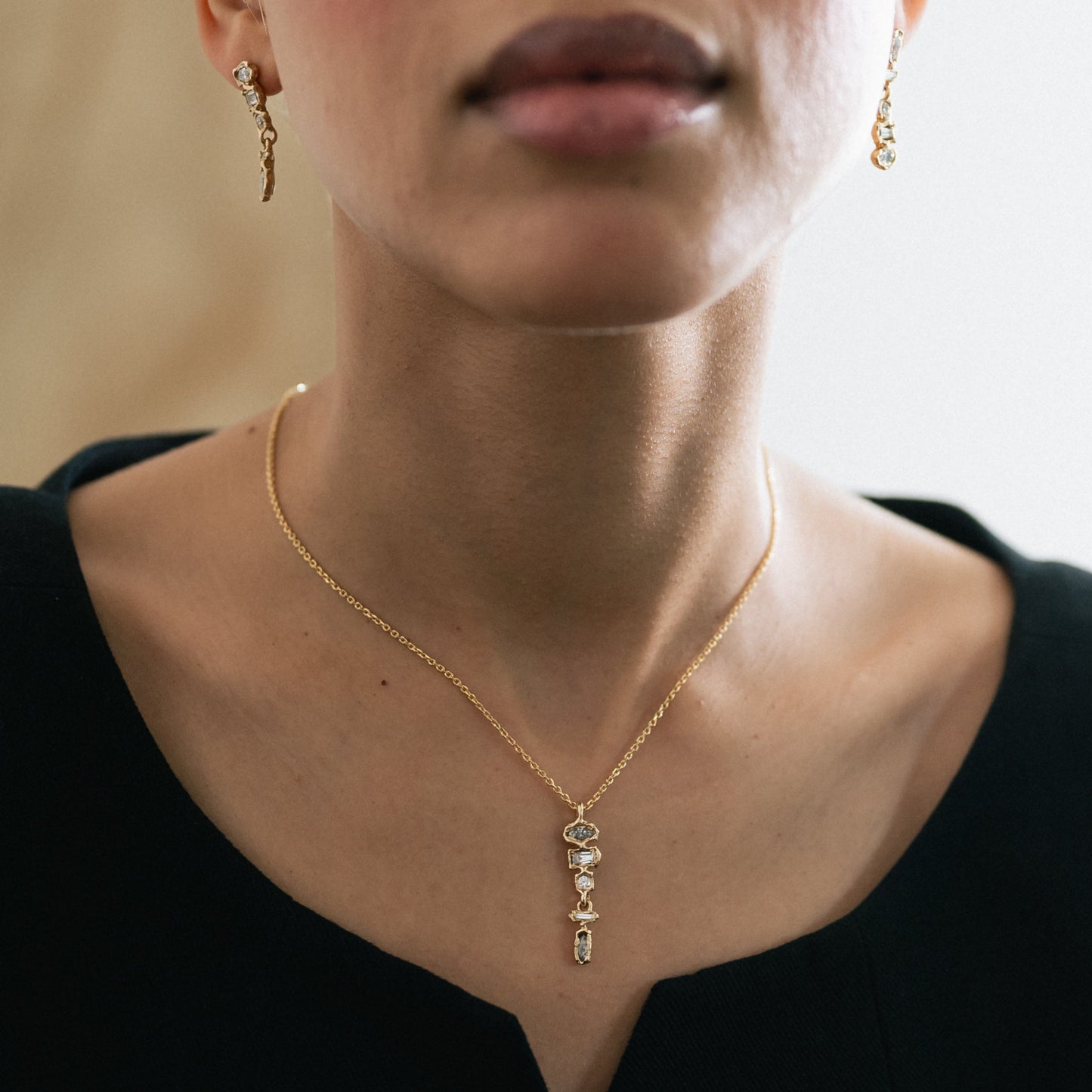 A close-up of a model wearing a delicate gold necklace with a small vertical pendant and matching dangling earrings, styled with a black top.