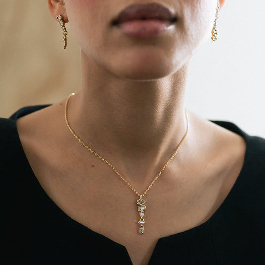 A close-up of a model wearing a delicate gold necklace with a small vertical pendant and matching dangling earrings, styled with a black top.