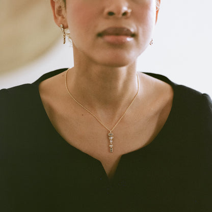 A close-up lifestyle shot of a model wearing a delicate gold key pendant necklace and matching key earrings, styled with a black V-neck top.