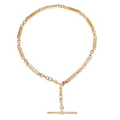 A gold-toned chain necklace with a unique link design featuring alternating cylindrical and rectangular segments, with a short hanging chain and a horizontal bar pendant at the bottom center, shown on a plain white background.