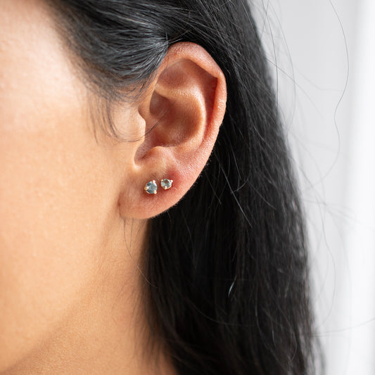 A close-up of a person's ear wearing two small gold stud earrings with blue stones, against a light neutral background.