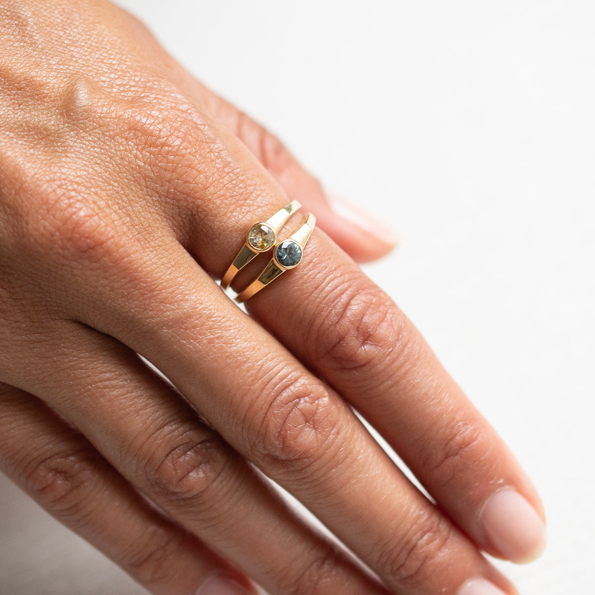 A close-up of a hand wearing two slim gold rings. One ring features a small round clear stone, and the other features a small round blue stone, set against a clean white background.