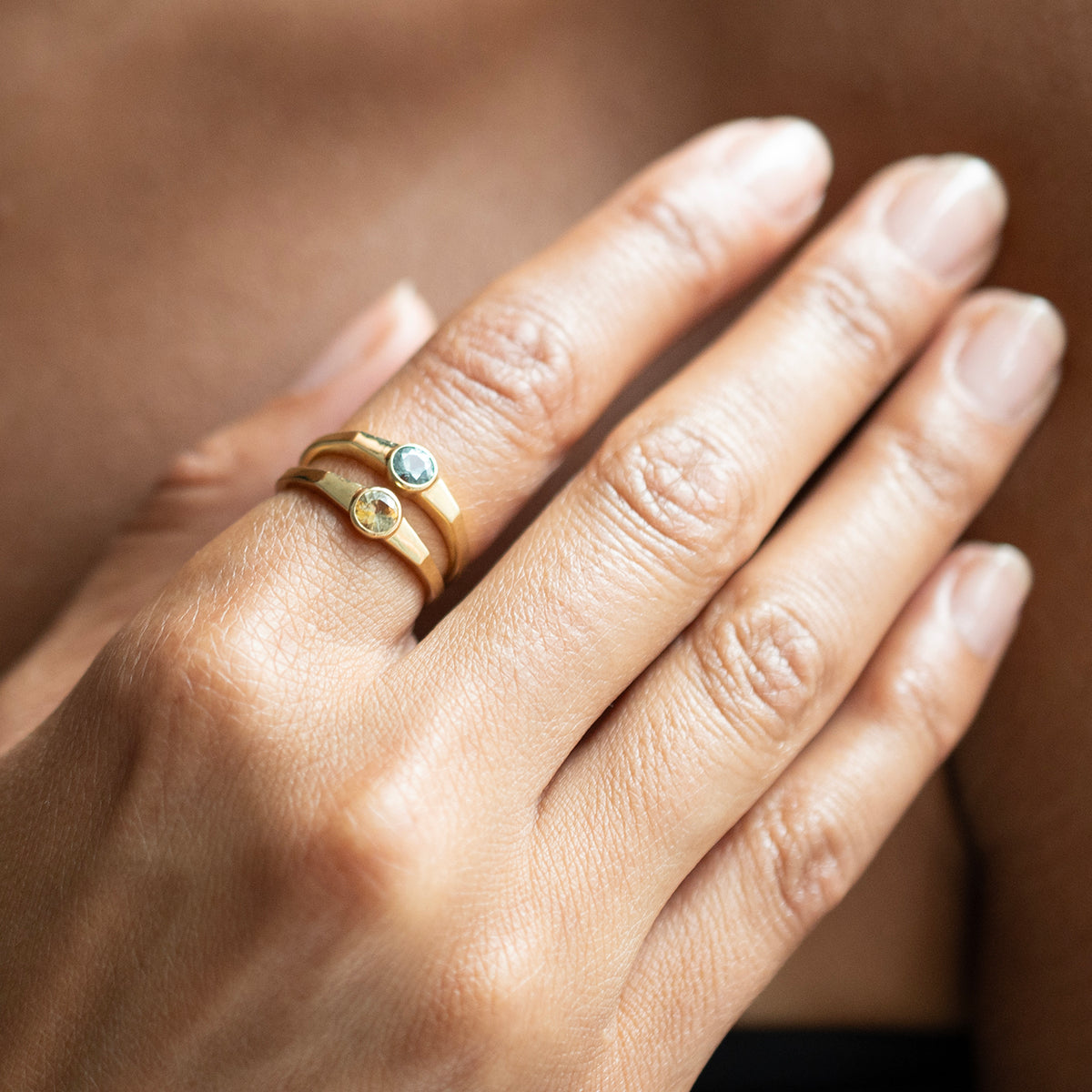 A close-up of a hand wearing two slim gold rings, one with a small round blue stone and the other with a small round yellow stone.
