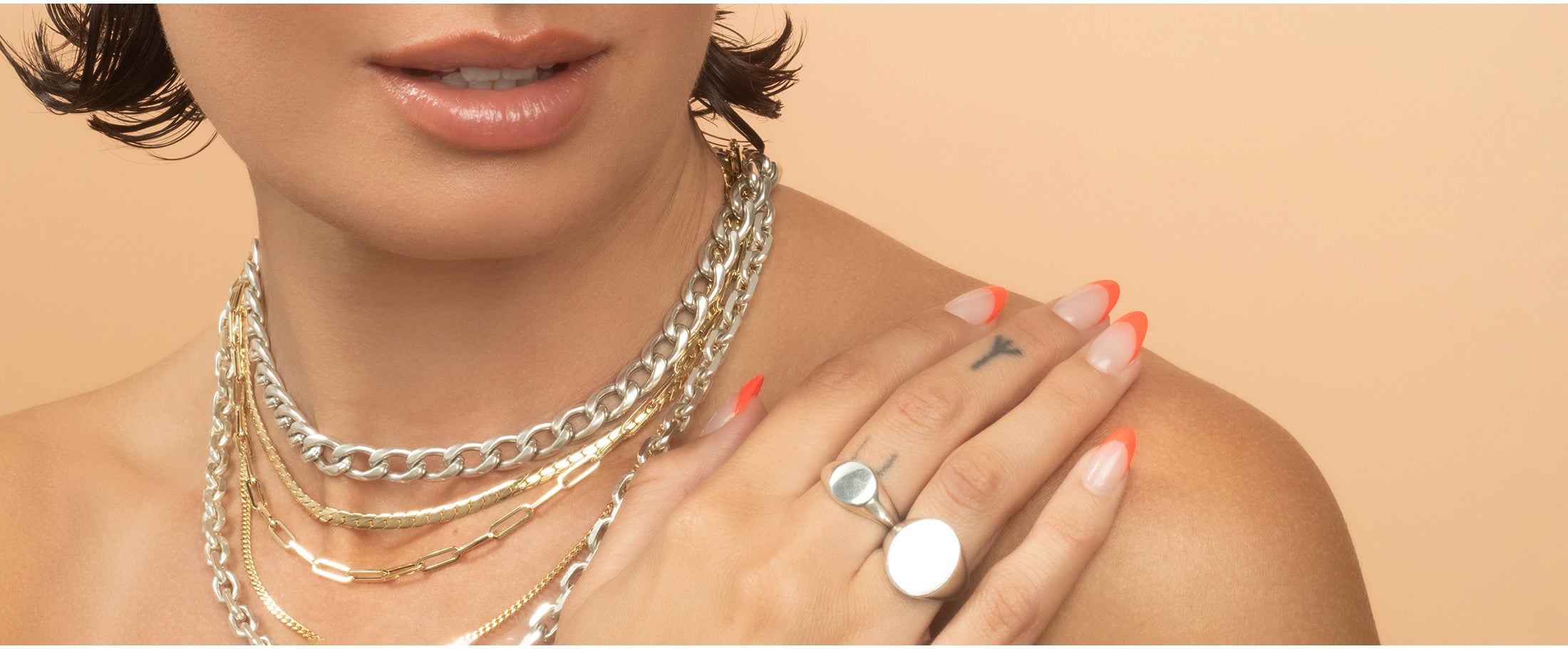 Close-up of a person wearing multiple gold and silver necklaces and silver rings on a beige background