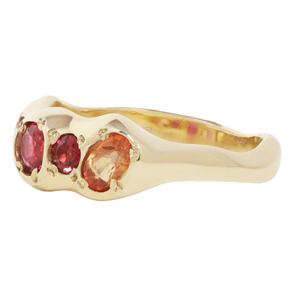 A yellow gold ring featuring three oval gemstones in pinkish-red and peachy-orange tones, set against a plain white background.