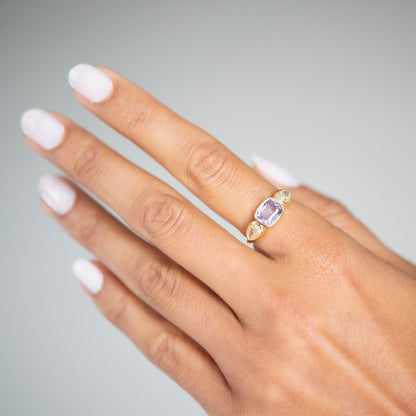 Lauren Wolf - Hand wearing a gold ring with a purple gemstone on a neutral background