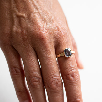 A close-up of a person's hand wearing a gold ring with a rectangular dark blue stone on a plain white background.