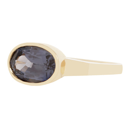 A gold ring featuring a large, faceted oval smoky blue gemstone in a bezel setting, shown against a plain white background.