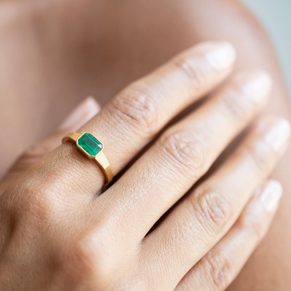 A close-up of a hand wearing a gold ring with a rectangular green emerald-cut gemstone.