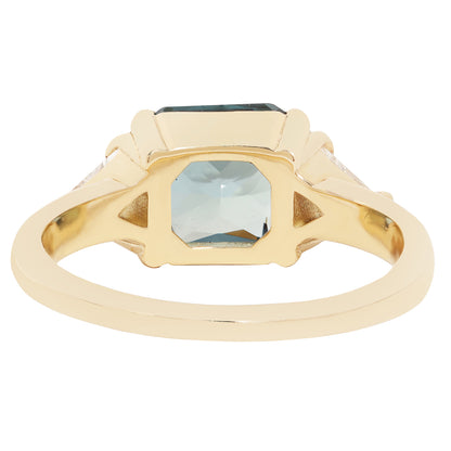 A gold signet ring featuring a large, faceted light blue-gray rectangular gemstone, with smaller clear stones set in the shoulders, shown on a plain white background.