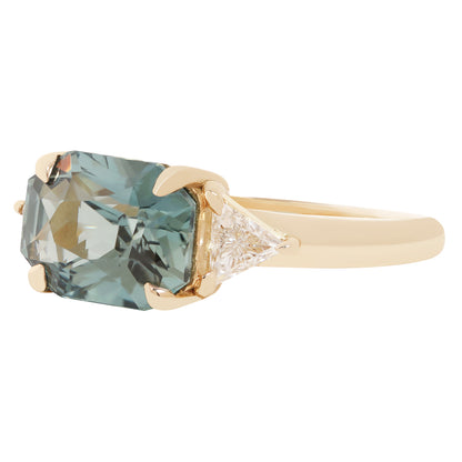 A yellow gold ring featuring a rectangular cushion-cut light blue gemstone and a small clear triangular accent stone on a white background.