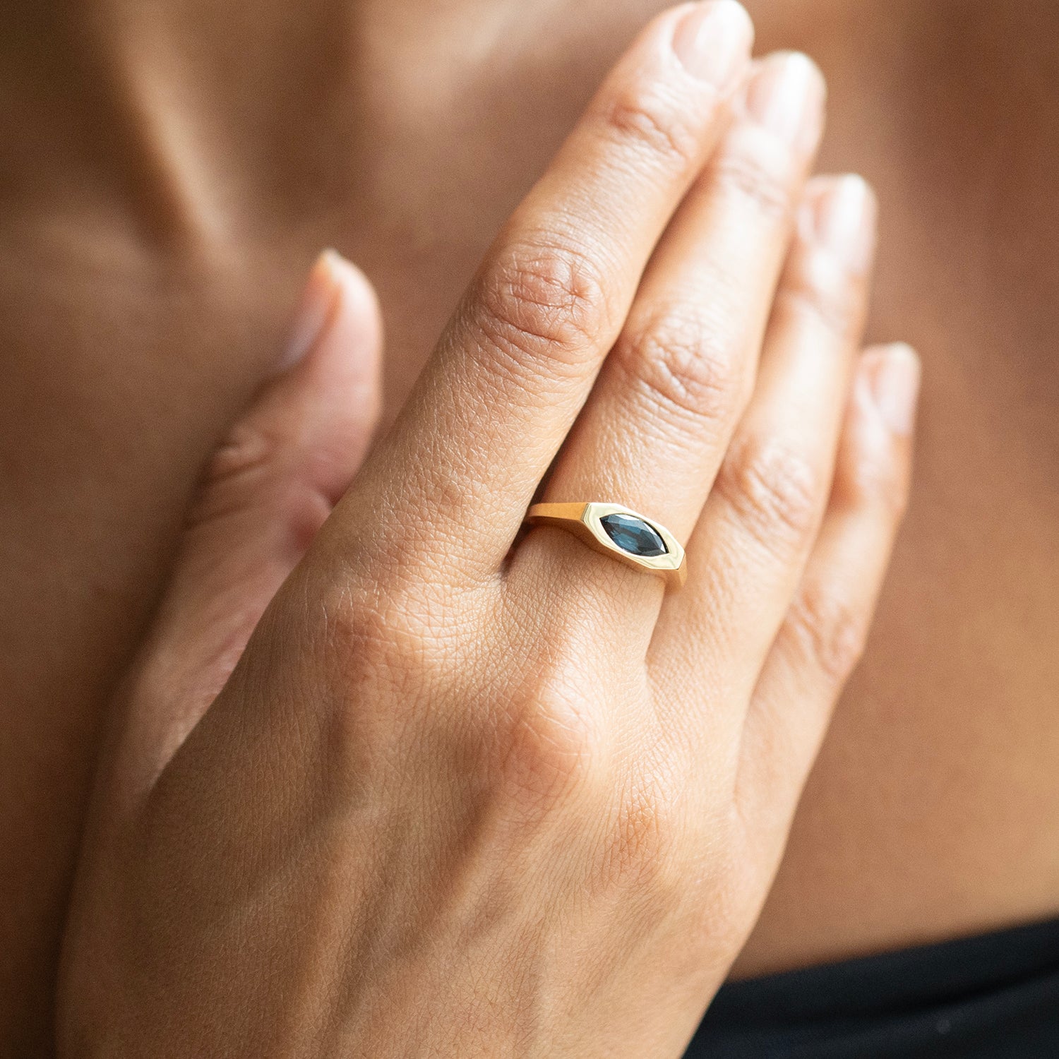 A close-up of a hand wearing a gold ring with a marquise-cut dark blue gemstone, set against a soft-focus background of skin.