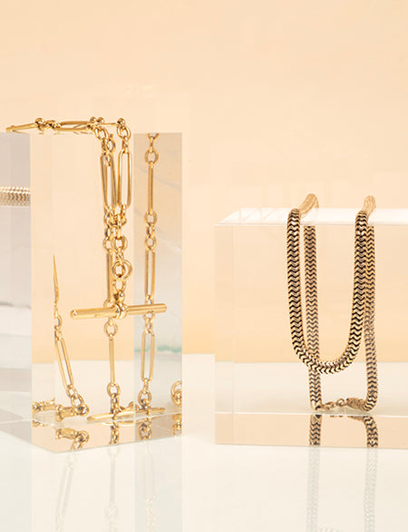 Two gold necklaces displayed on clear stands against a beige background
