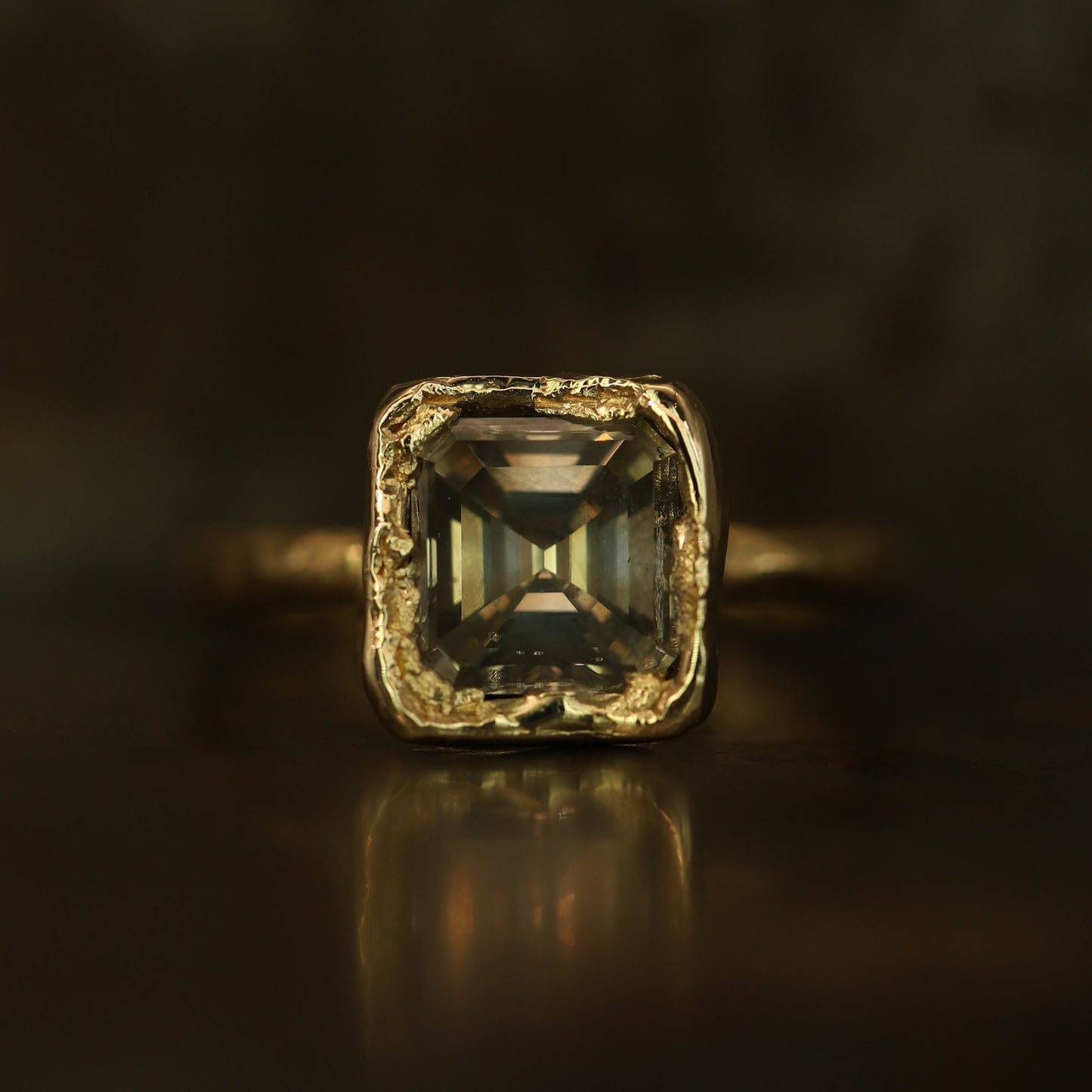 A square emerald-cut gemstone ring in a textured gold setting, resting on a dark reflective surface with a soft reflection.