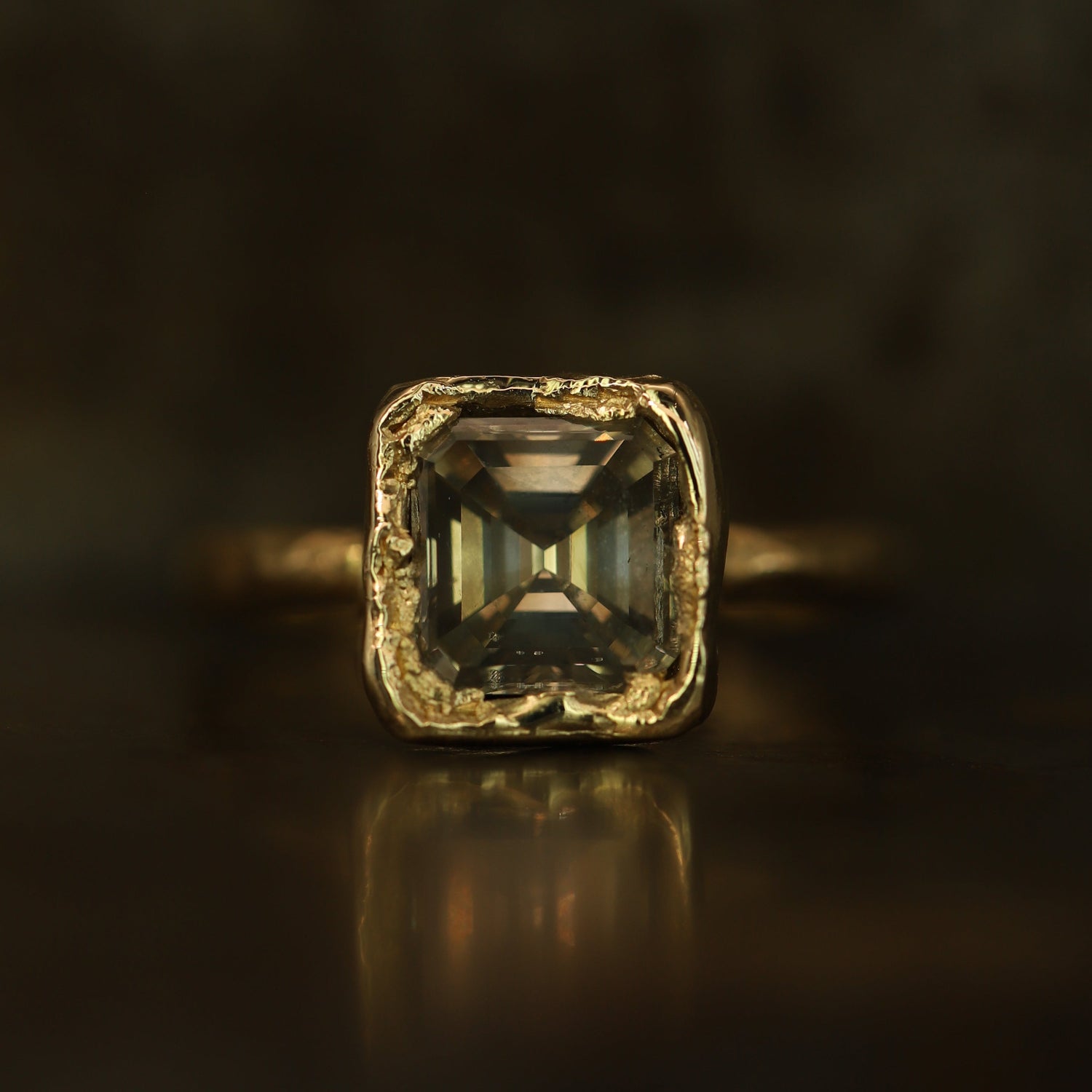A square emerald-cut gemstone ring in a textured gold setting, resting on a dark reflective surface with a soft reflection.