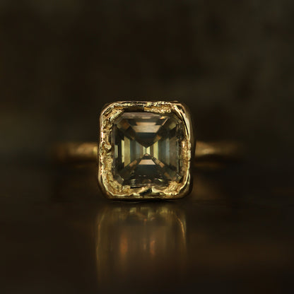 A square emerald-cut gemstone ring in a textured gold setting, resting on a dark reflective surface with a soft reflection.