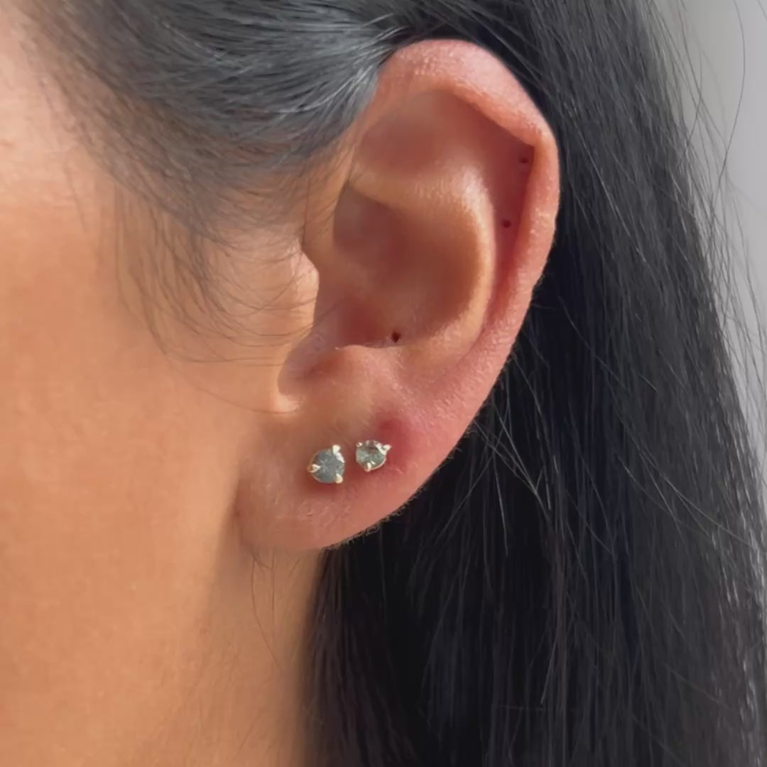 A close-up of a person's ear wearing two small gold stud earrings with blue stones, against a light neutral background.