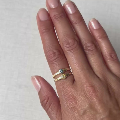 A close-up of a hand wearing a delicate gold ring with a small round sapphire on a plain white background.
